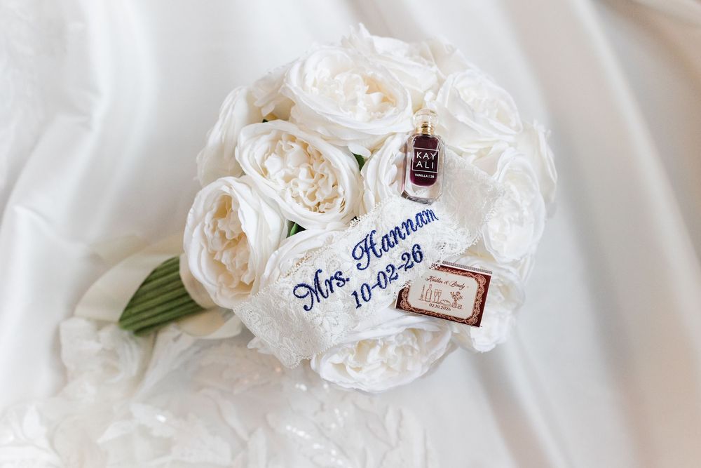 White rose wedding bouquet with a blue embroidered Mrs. Hannam 10-02-26 ribbon and small framed photo charms on a white satin background.