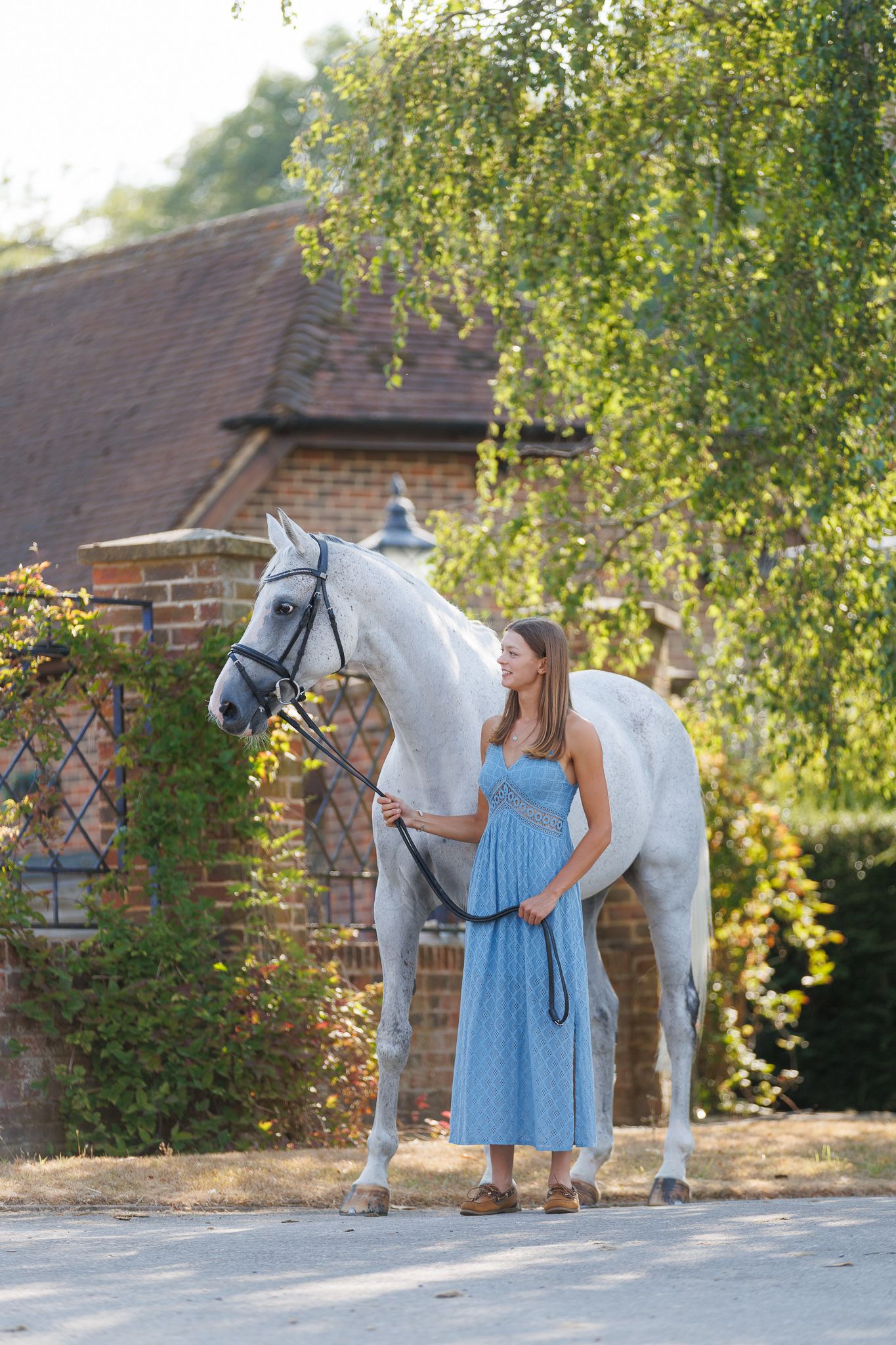 Woman in a blue sundress stands holding the lead rope of a gray horse in a bridle beside a brick building and green trees.