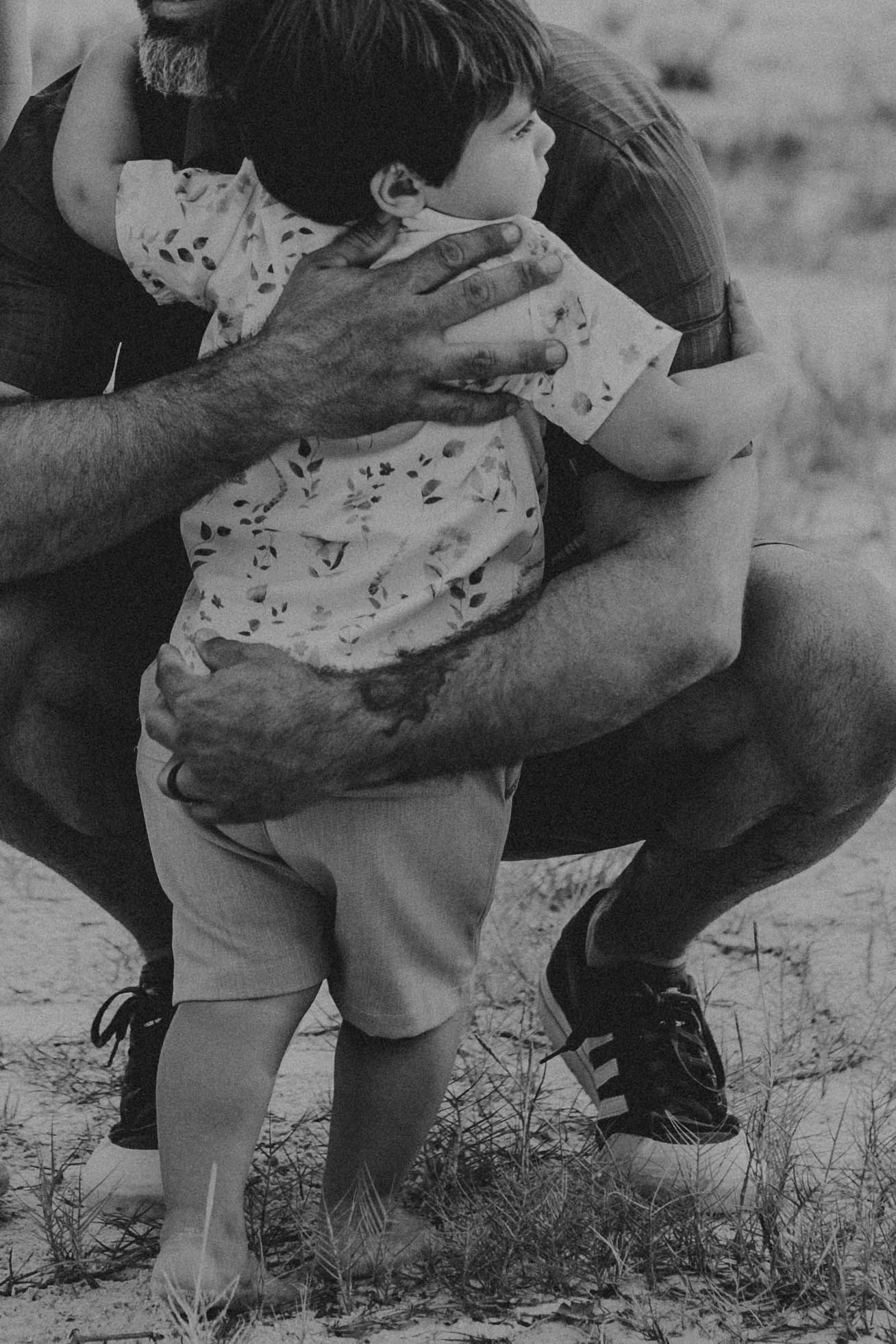 Black and white photo of a tattooed man crouching outdoors and hugging a toddler in a floral shirt standing barefoot on grassy sand.