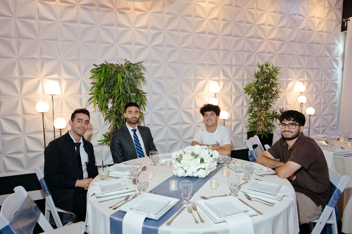 Four men sit around a round banquet table set with white linens, plates and glasses in an elegant room with white geometric wall panels.
