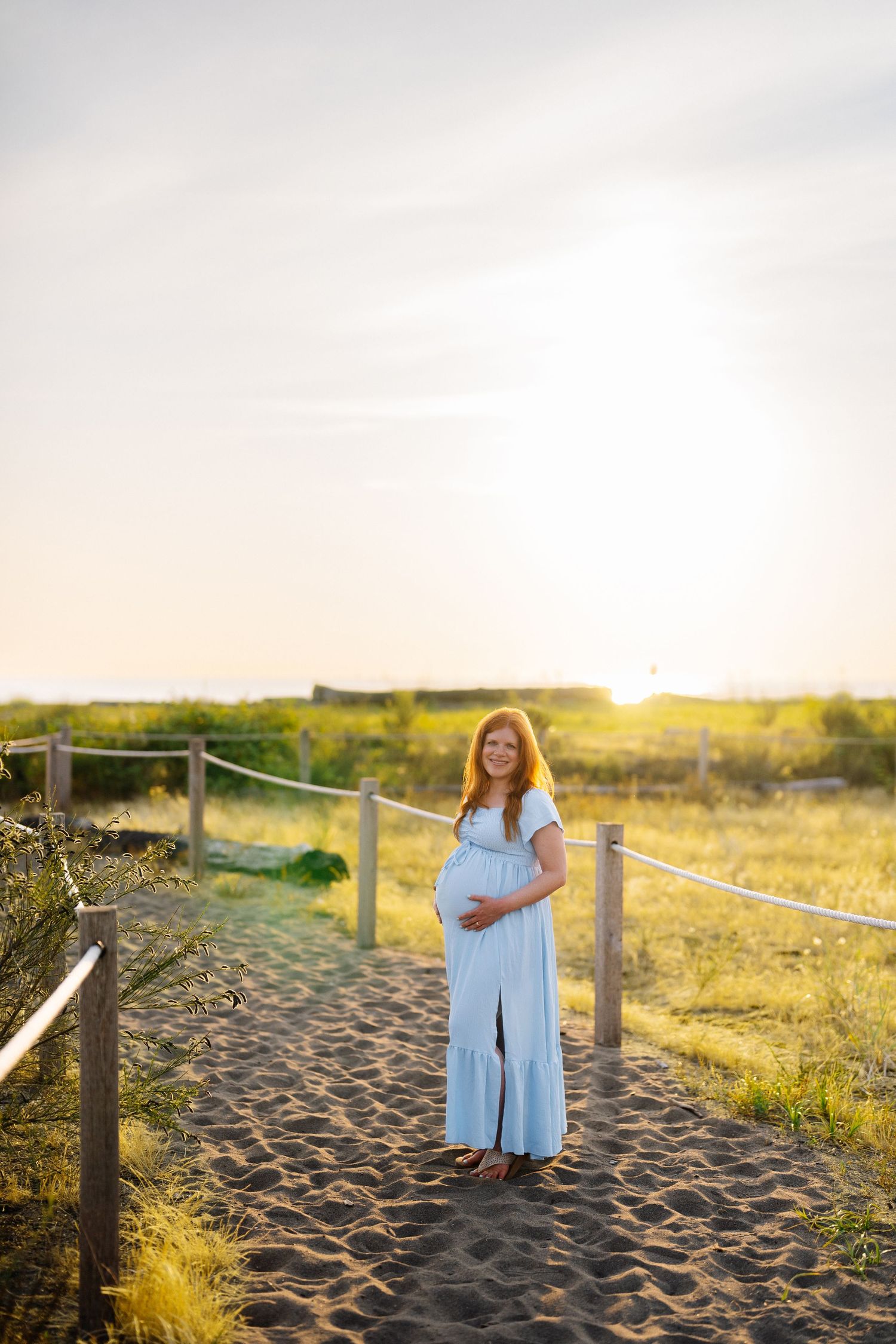 Pregnant woman in a flowing light blue dress cradles her belly on a sandy beach path at golden hour sunset.