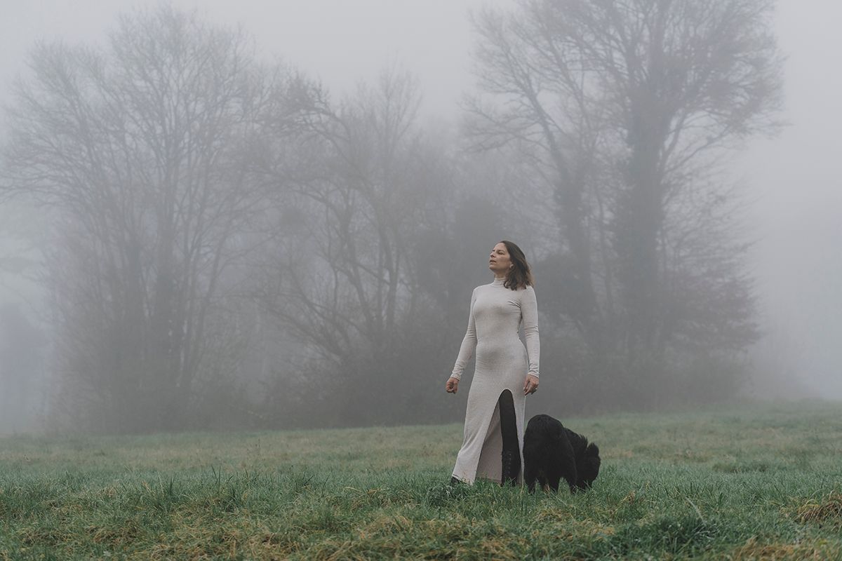 Woman in a long white dress stands in a misty green field with a black dog, surrounded by foggy bare trees.