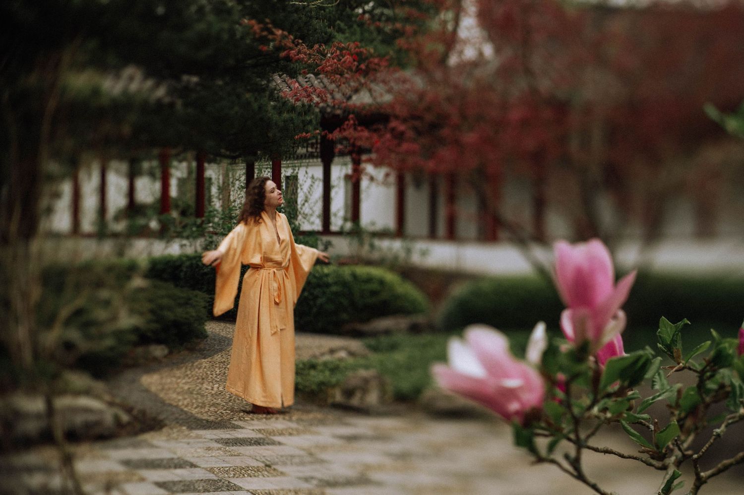 Woman in a flowing golden robe walks along a stone path in a serene Japanese botanic garden with pink blossoms in the foreground.