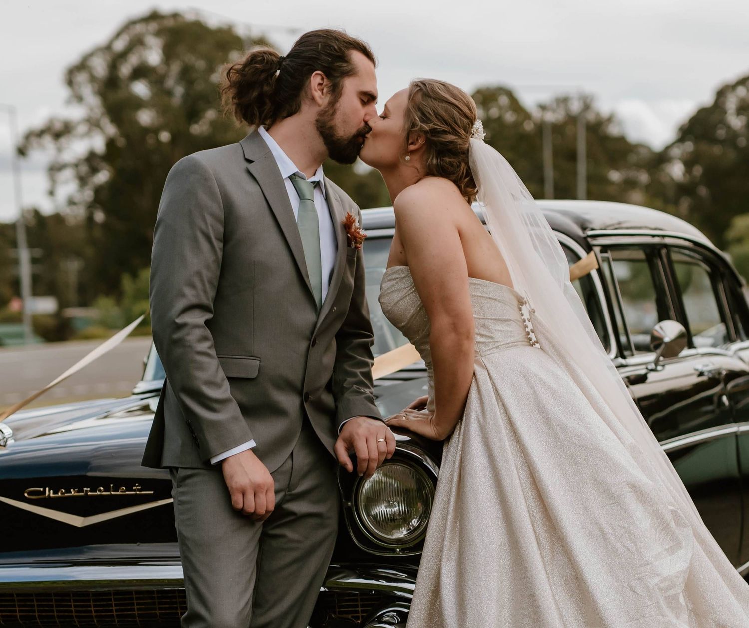 Bride and groom kissing in front of a vintage black Chevrolet car, groom in gray suit, bride in white gown with veil.