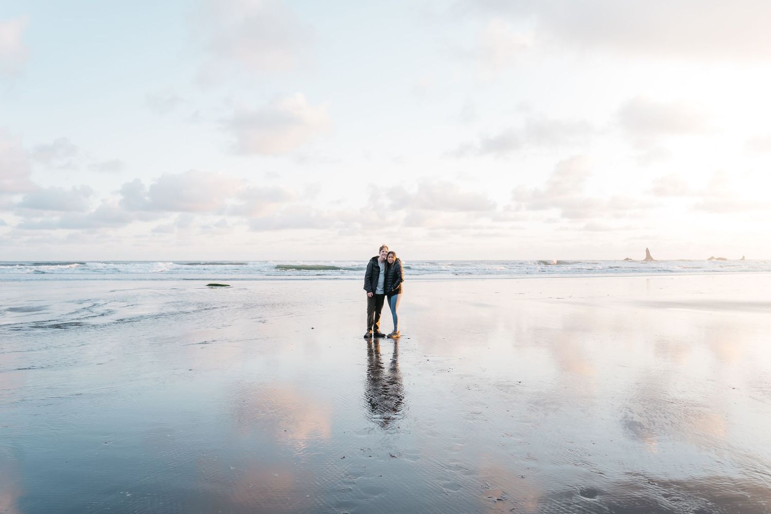 Couple embracing on a reflective wet beach at sunset with gentle waves and a lighthouse in the distance.