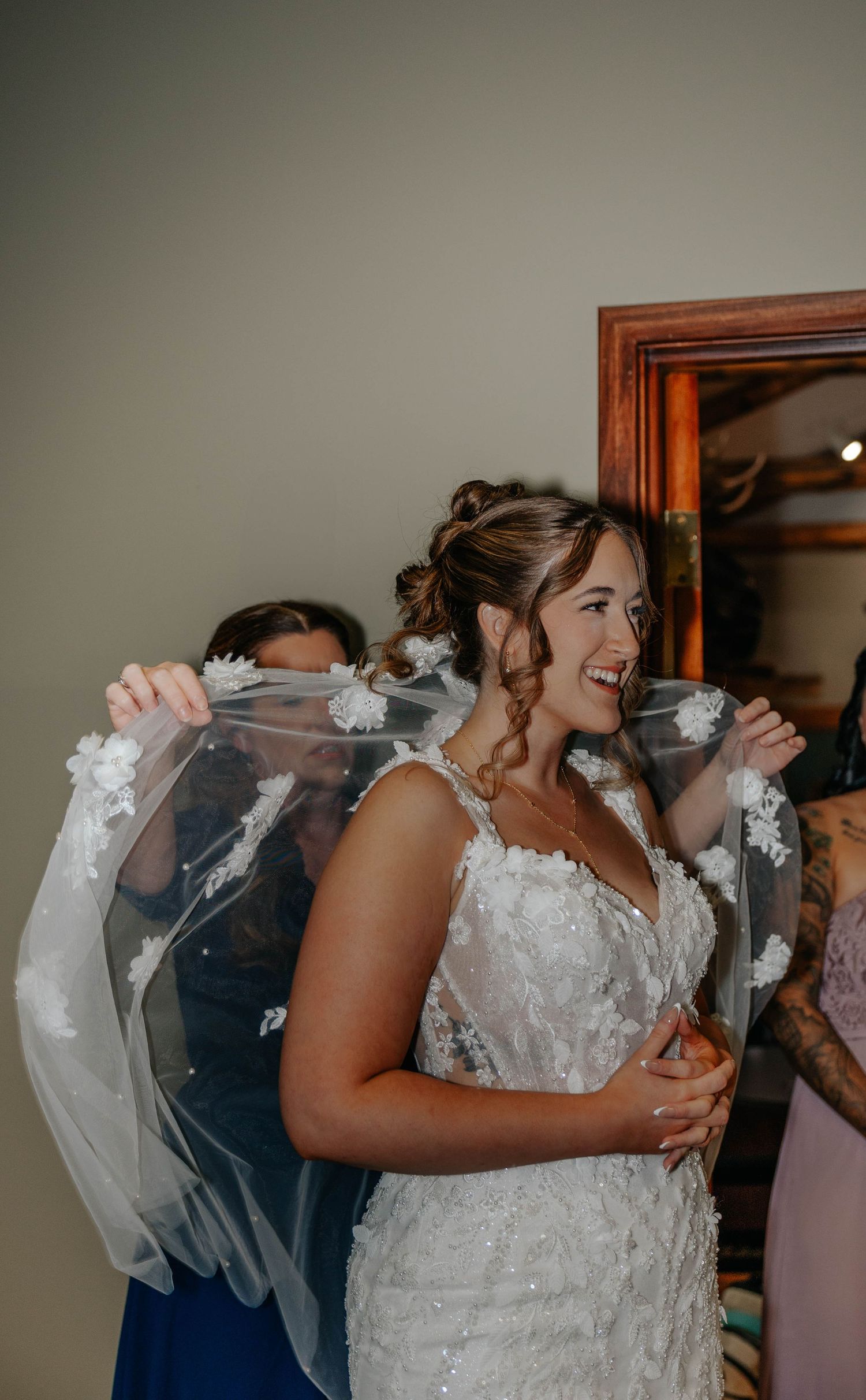 Smiling bride in lace wedding dress having veil placed by bridesmaids before ceremony.