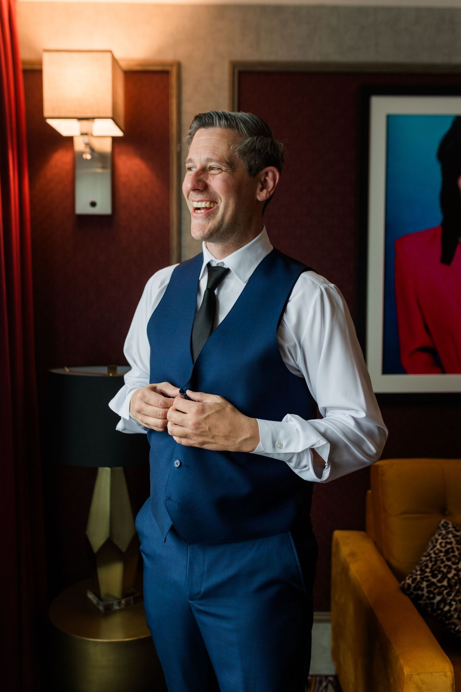 Smiling man in a blue vest, white shirt and dark tie adjusts his cuffs while getting ready in a warmly lit room with a colorful portrait.