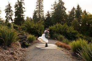 Couple sharing a romantic dip kiss on a garden path surrounded by lush greenery and tall evergreen trees. Captured by Savvy Photography