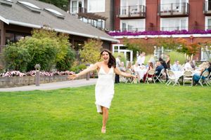 Bride in white dress running joyfully on green lawn at outdoor wedding reception with guests seated in background. Captured by Savvy Photogr