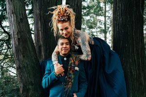 Two people smiling and embracing outdoors among tall trees, one with dreadlocks and tattoos wearing a floral outfit.