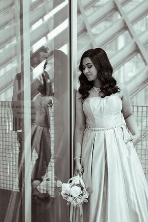 Black and white photo of a bride in a strapless gown holding a bouquet, with a groom reflected in glass behind her. Captured by Savvy