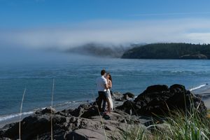 Couple embracing on rocky Pacific coast shoreline with ocean waves, green hills, and dramatic fog rolling in behind them. Captured by Savvy