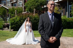 A bride in a white gown with veil stands behind her dad in a gray suit during an outdoor first look wedding moment. Captured by Savvy