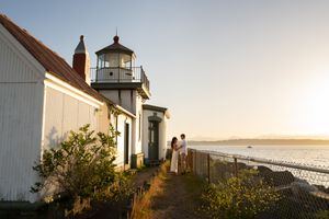 Couple embracing near a white lighthouse at golden hour with ocean views in the background.