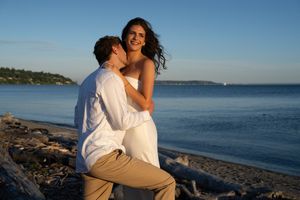 Couple sharing a romantic moment on a rocky beach at sunset, woman in white dress smiling joyfully.