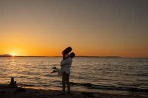 Couple embracing at sunset on a beach with golden sky and calm ocean water in the background.