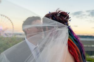 Bride with rainbow hair kissing groom under veil at sunset with Ferris wheel in background. Captured by Savvy Photography