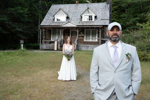 Bride in white gown holds bouquet behind groom in gray suit and cap outside rustic log cabin wedding venue. Captured by Savvy Photography