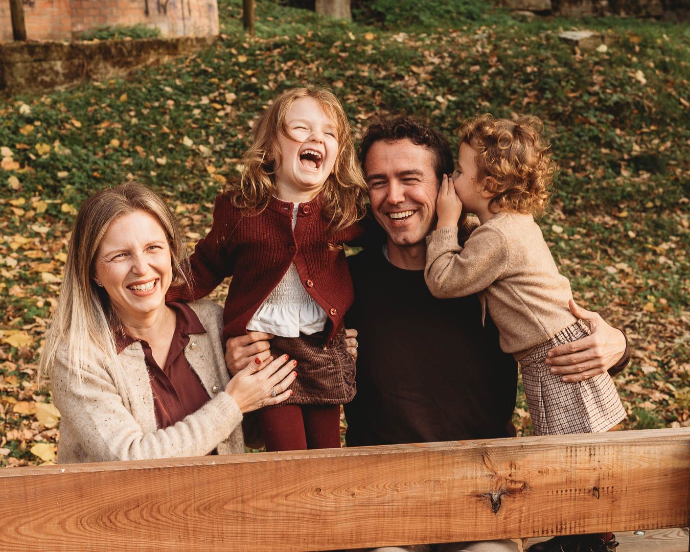 Happy family of four laughing outdoors in autumn park, parents holding two young children near wooden fence.