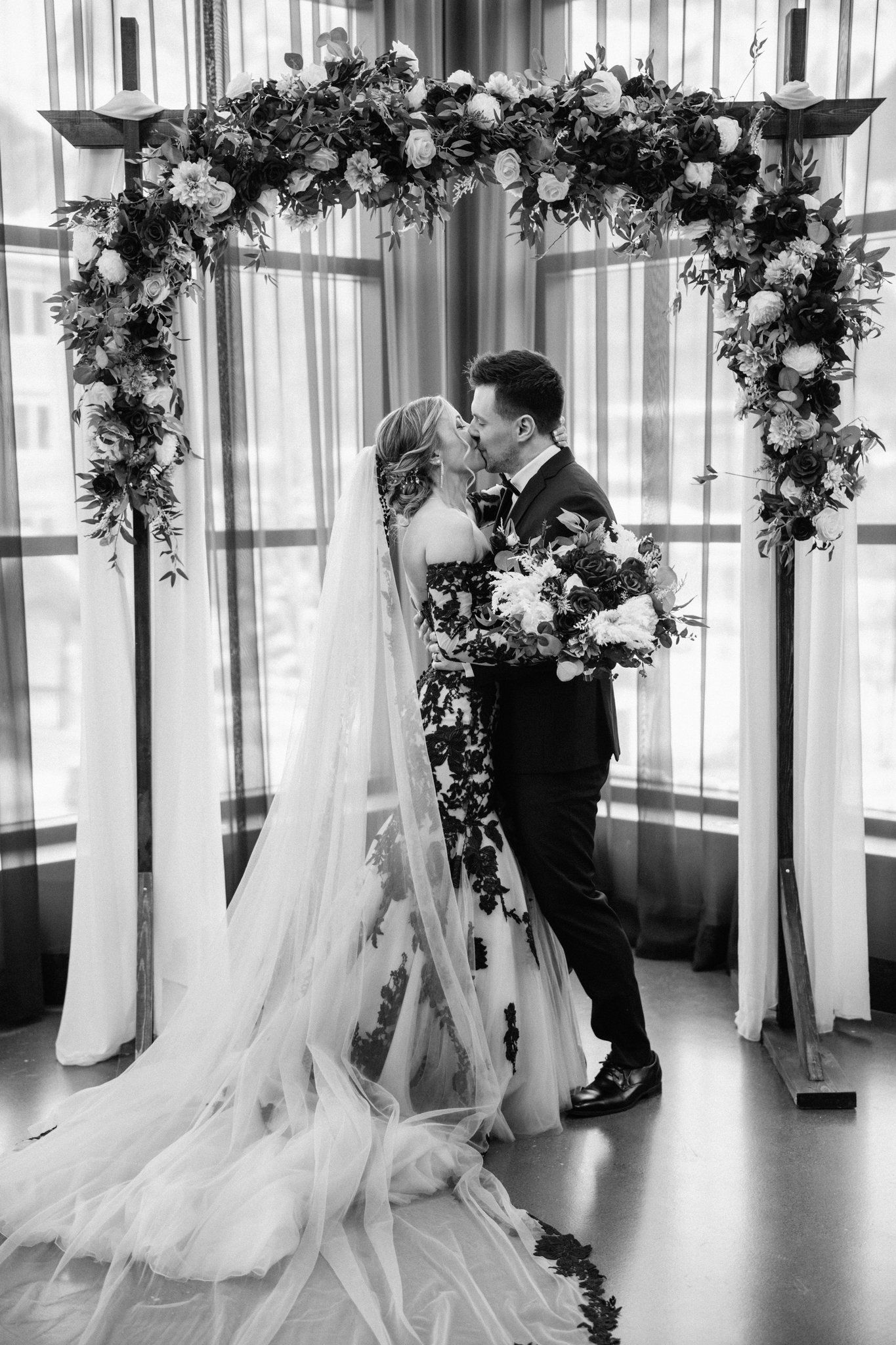 Black and white first kiss under a floral arch in front of tall windows at Mainspace