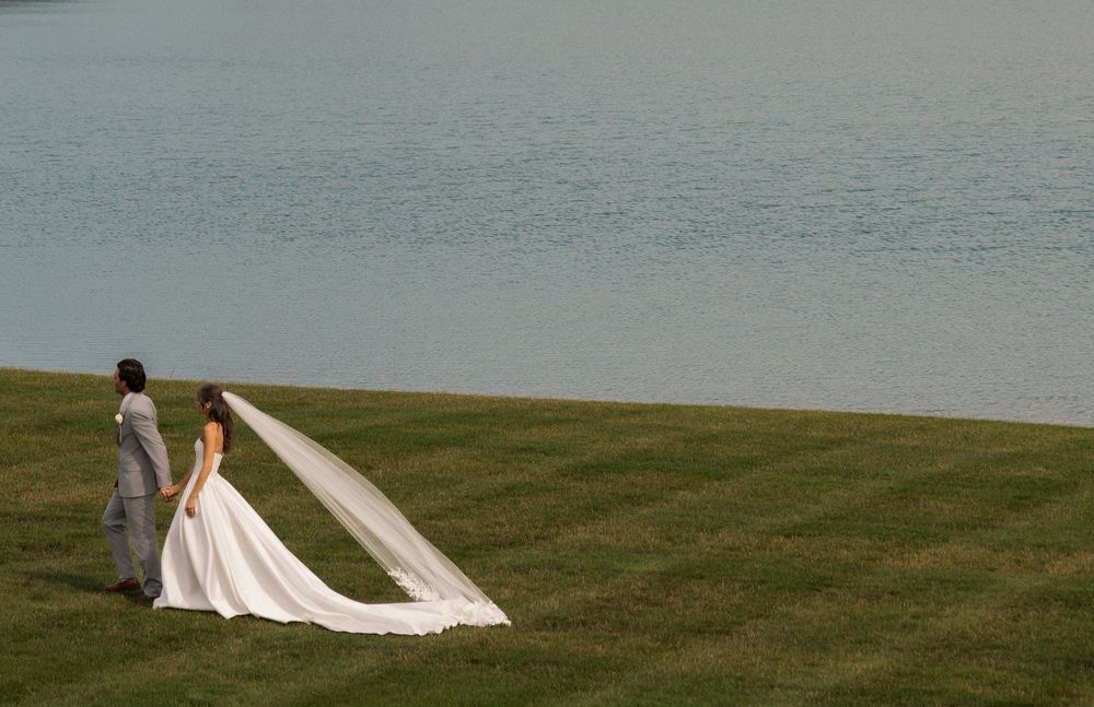 Bride and groom holding hands on green lawn by serene lake, bride wearing elegant white gown with long flowing veil.