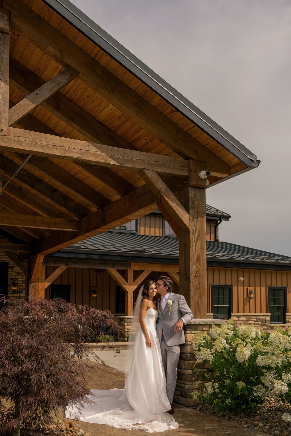 Bride and groom pose outside a rustic wooden wedding venue with stone accents and lush greenery under an overcast sky.