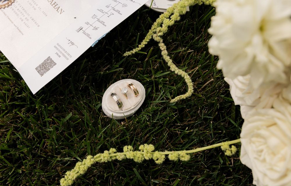 Wedding rings in a decorative holder on green grass beside white flowers and a marriage certificate.