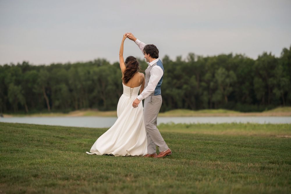 Bride and groom dancing outdoors near a lake, surrounded by lush green trees on their wedding day.