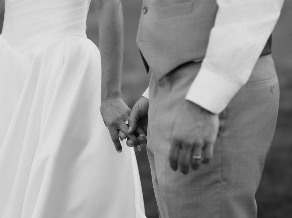 Black and white photo of bride and groom holding hands, showcasing wedding rings at their elegant ceremony.