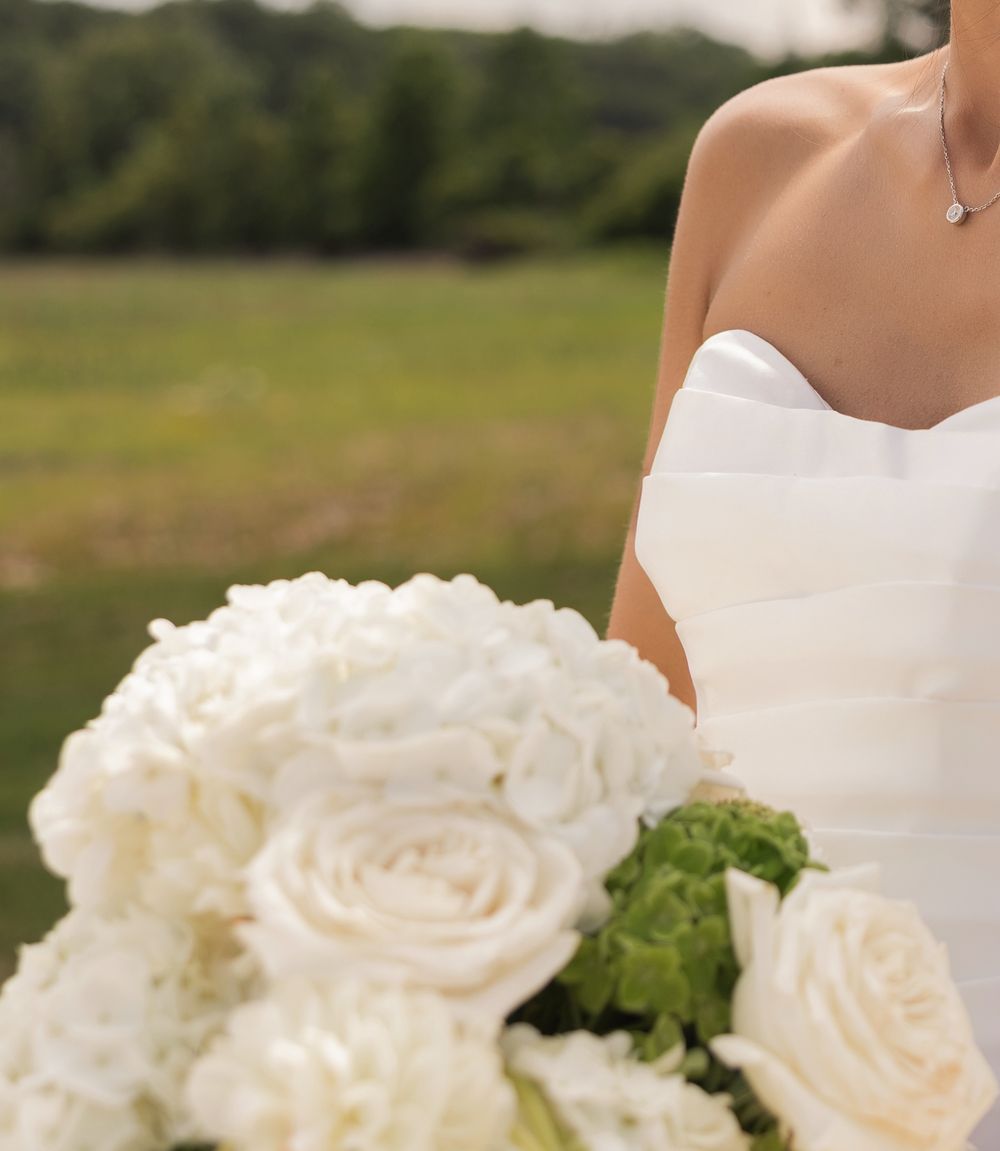 Bride in white strapless gown holding elegant bouquet of white roses and hydrangeas outdoors.