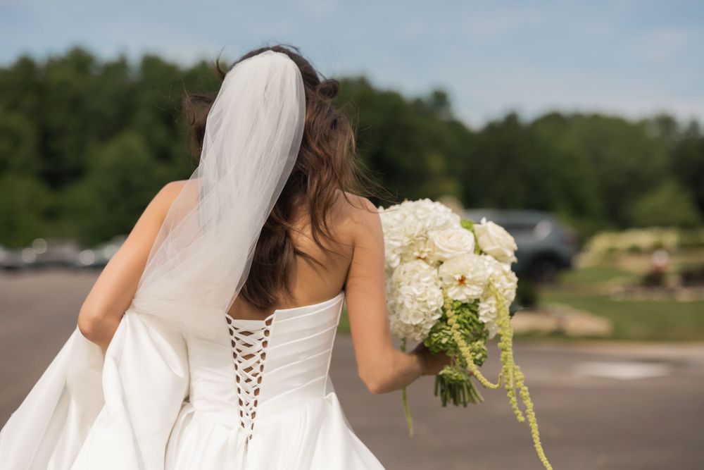 Bride in white strapless corset gown holding white bouquet, veil flowing, standing outdoors with trees in background.