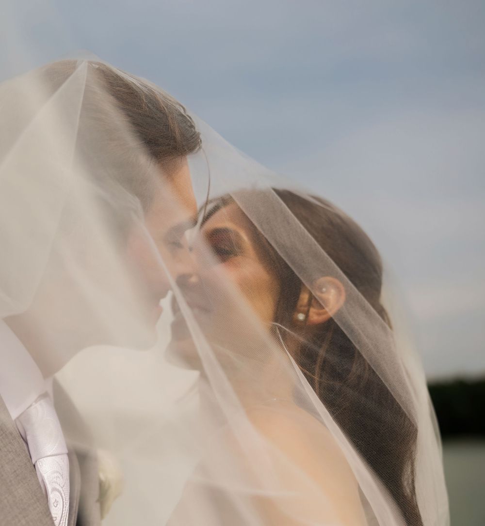 Bride and groom sharing a romantic kiss under a flowing white wedding veil outdoors on their wedding day.