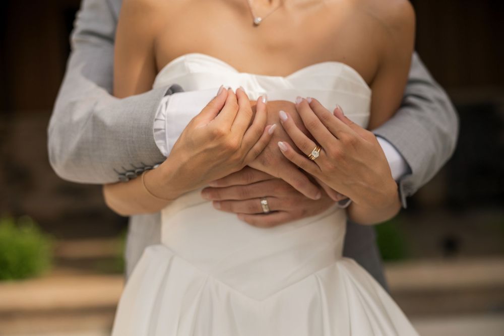 Bride and groom embracing, showing wedding rings on clasped hands, white bridal gown and gray suit.