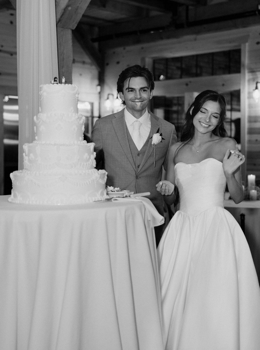 Bride and groom smiling beside a tall white wedding cake in a rustic venue, black and white photo.