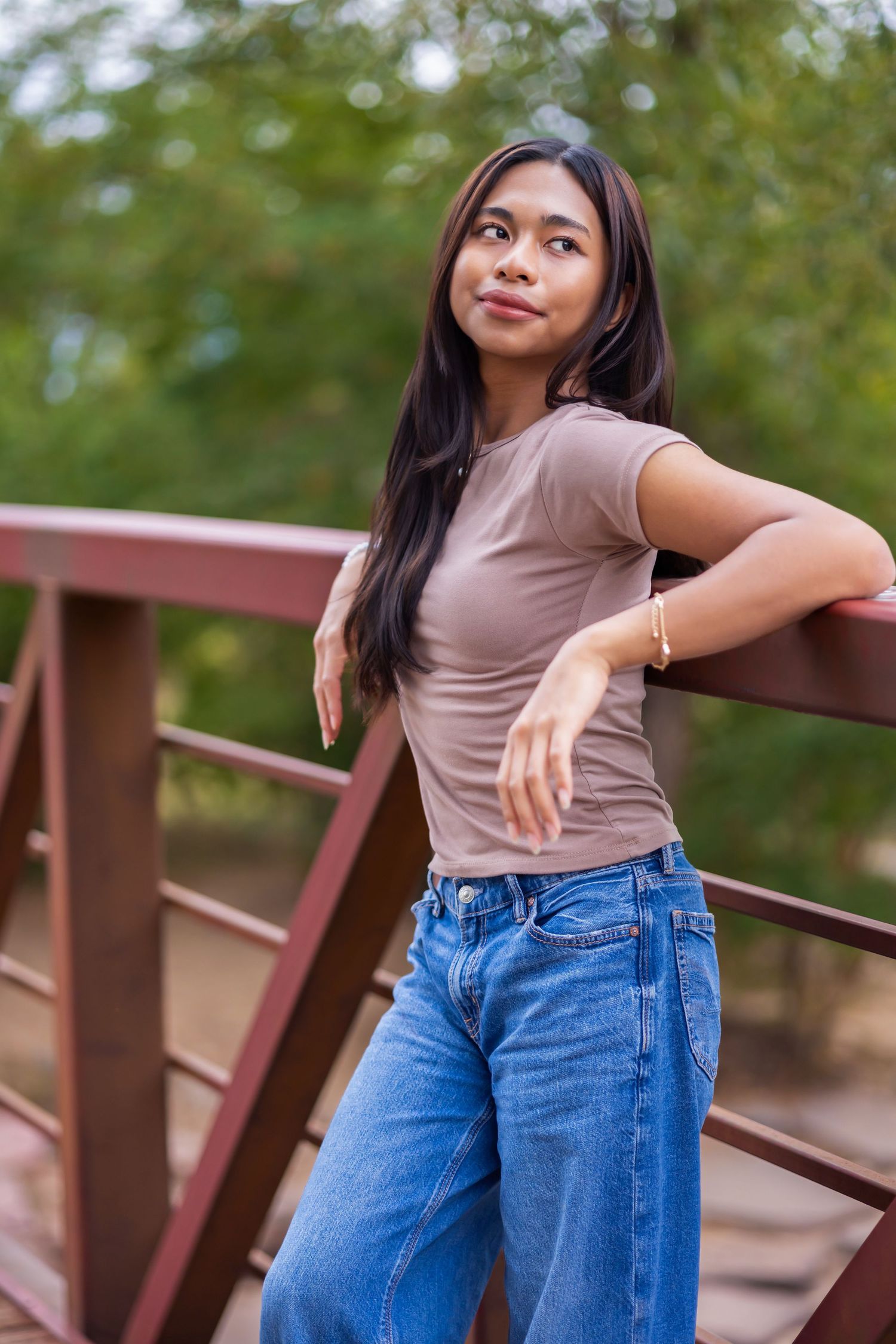 Young Asian woman in brown tee and blue jeans posing on a red metal bridge surrounded by green trees.