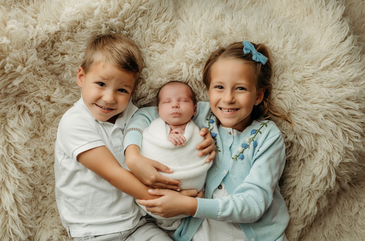 Two young siblings lying on a fluffy rug, smiling and holding a swaddled newborn baby between them.
