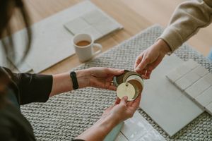Two people exchanging a small wooden tag over a textured table with tea and tiles in a cozy setting.