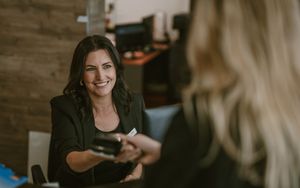 Smiling brunette woman at a reception desk handing a card reader to a blonde customer for payment.