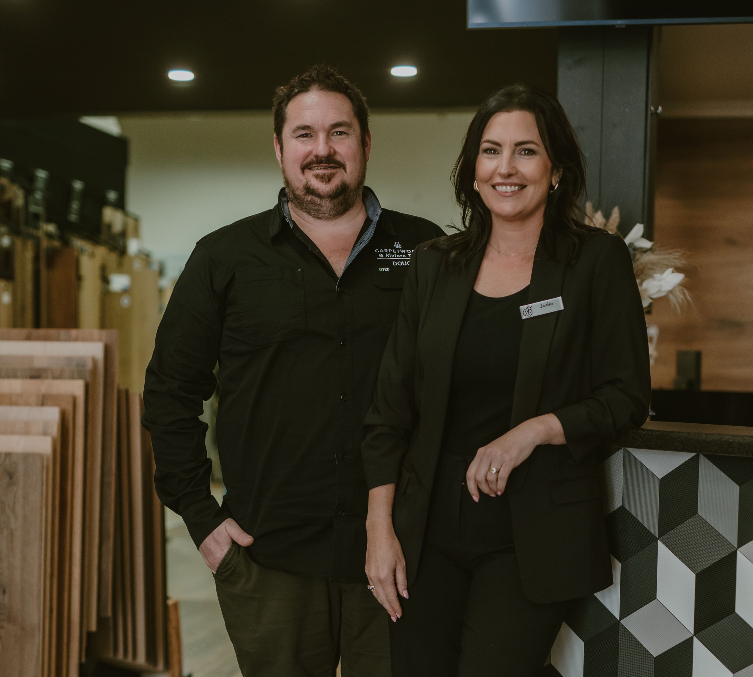 Two smiling professionals in black attire pose together in a modern flooring showroom.