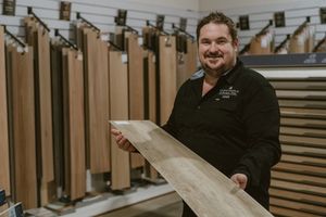 Man smiling in flooring showroom holding a wood plank sample surrounded by display racks of hardwood flooring options.