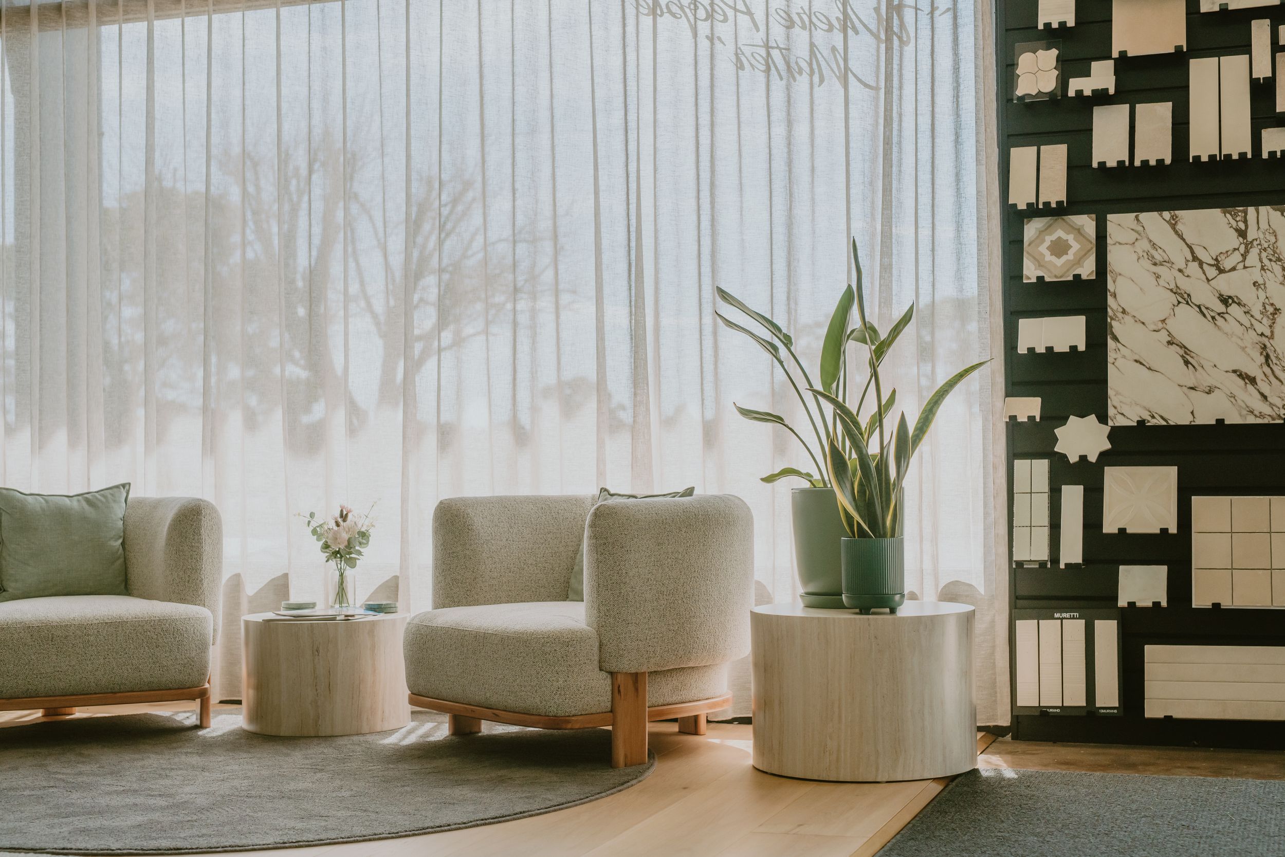 Modern minimalist lounge with white bouclé armchairs, sheer curtains, and a tropical plant near a large window.