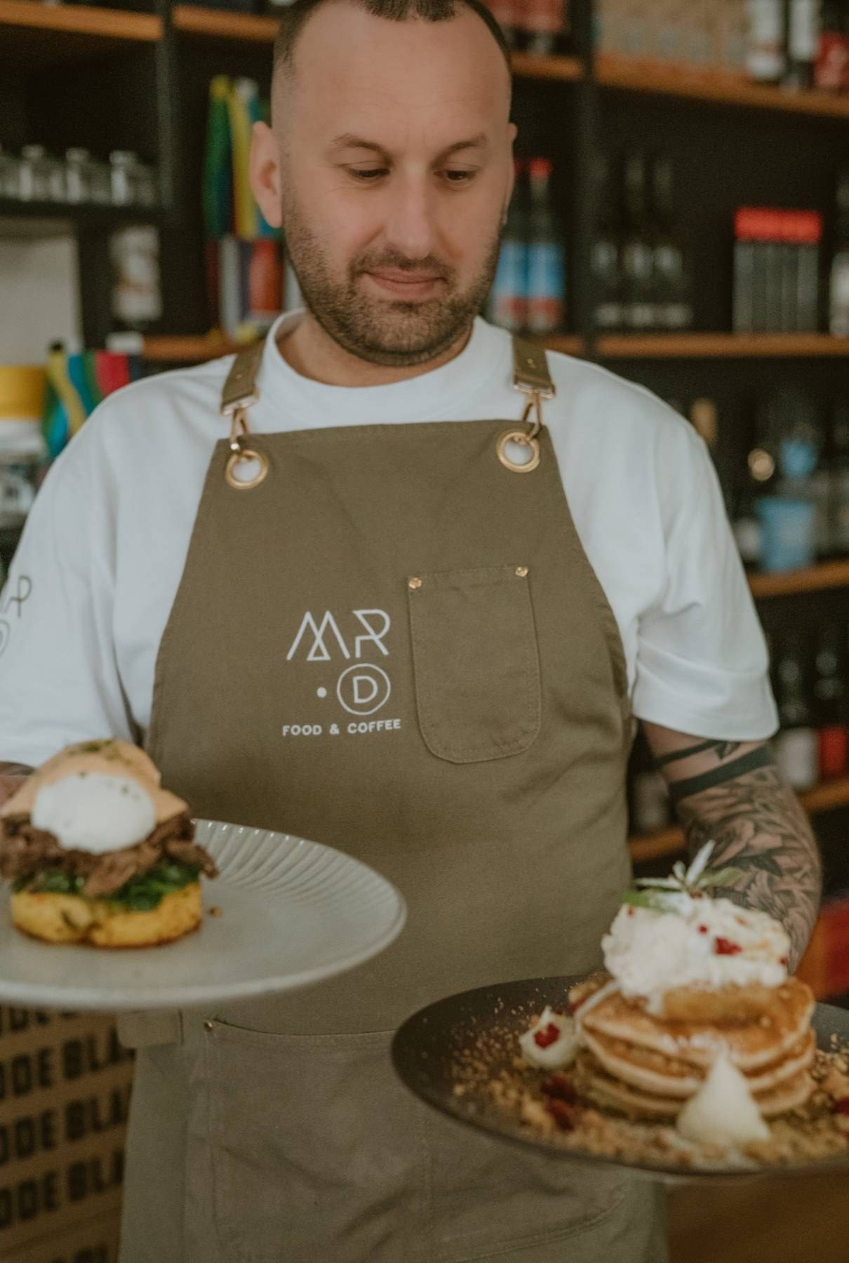 Tattooed chef in olive Mr. D Food & Coffee apron holds two plates of gourmet brunch dishes in a cafe.