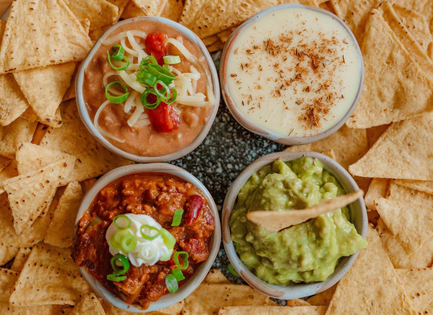 Four bowls of Mexican dips including guacamole, queso, chili, and bean dip surrounded by tortilla chips.