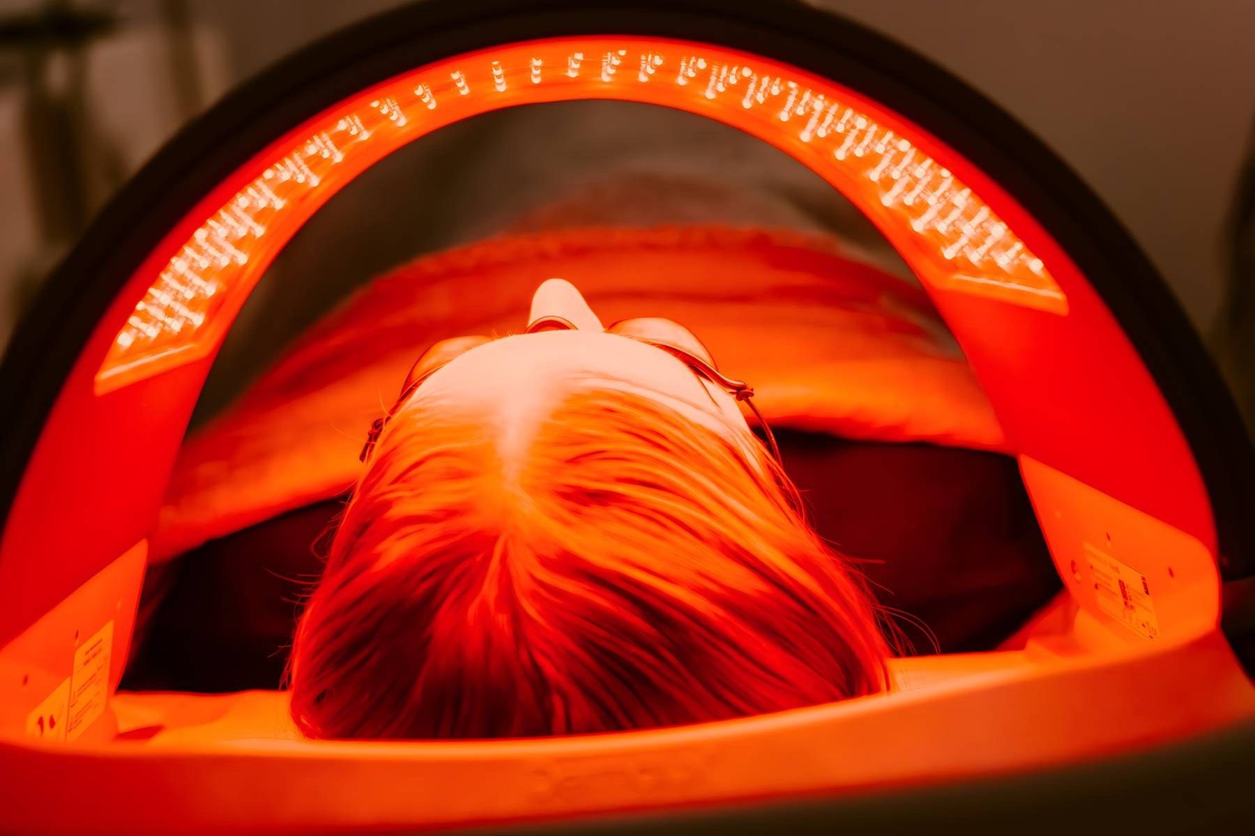 Person receiving red light therapy facial treatment under glowing LED arch device in a spa setting.