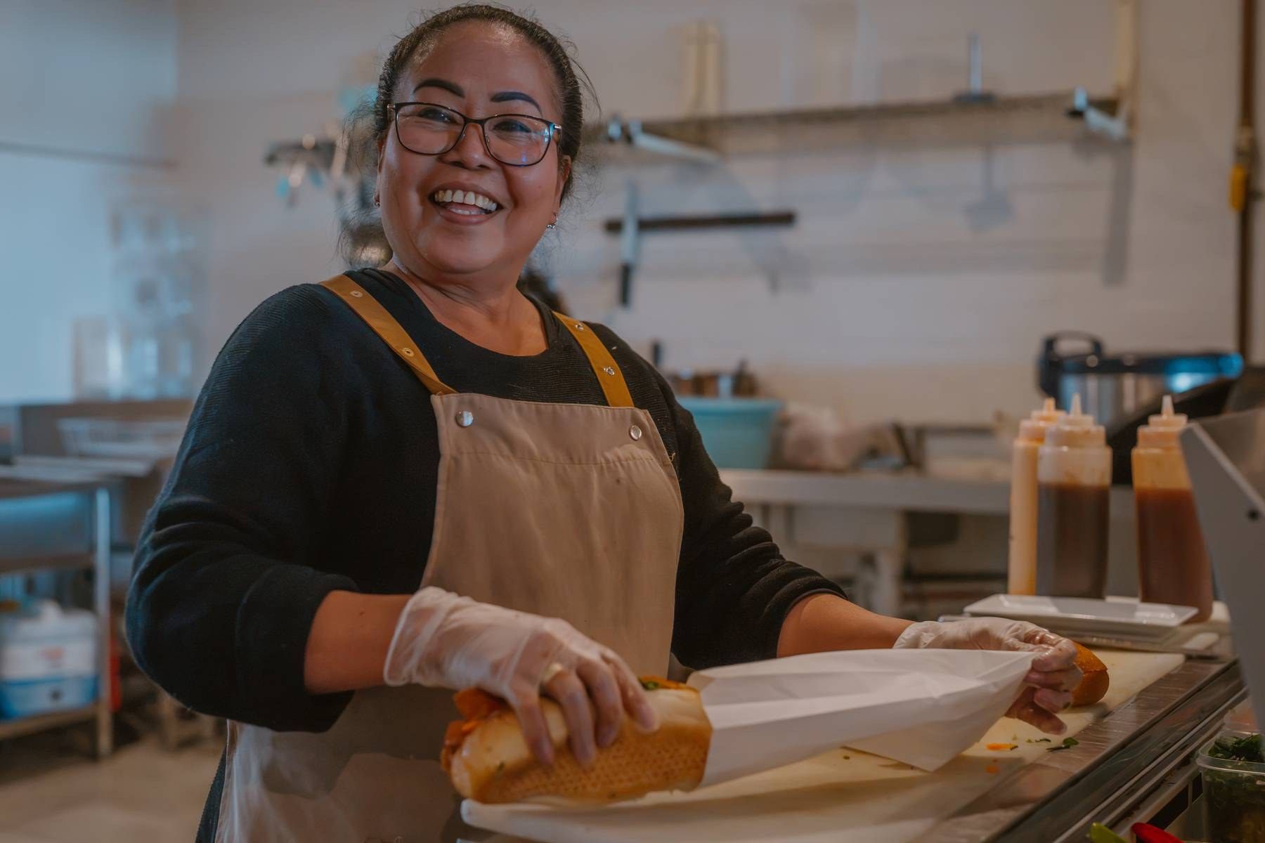 Smiling woman in apron prepares a sandwich in a commercial kitchen, wearing gloves and glasses.