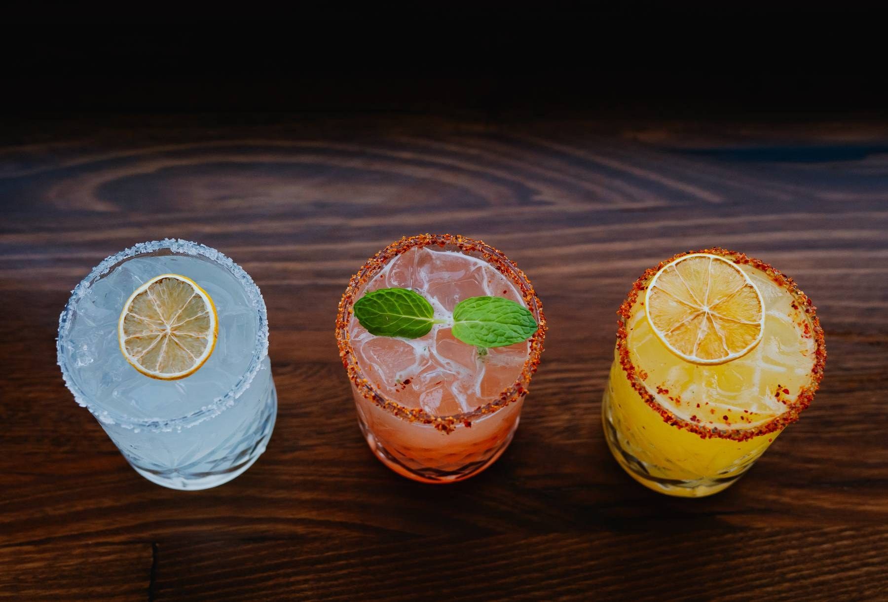 Three colorful margaritas with salted rims, garnished with citrus slices and mint, served on a rustic wooden table.