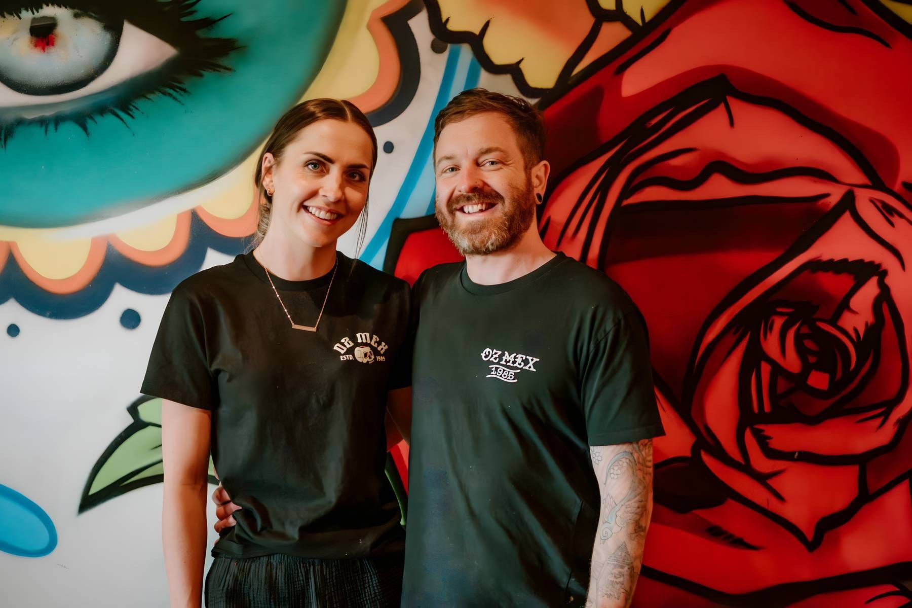 Two smiling people in black t-shirts pose in front of a vibrant colorful graffiti mural with roses.