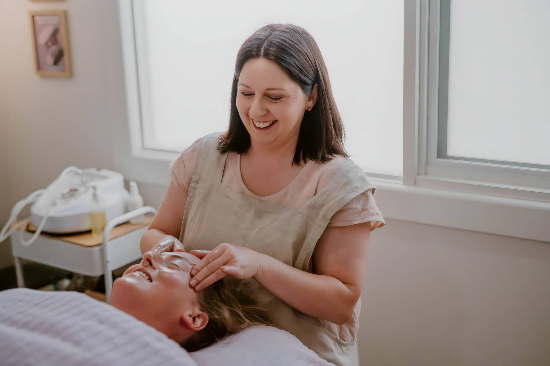 Smiling esthetician performing a relaxing facial treatment on a client in a bright, modern spa room.