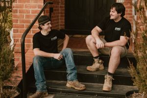 Two men in matching black shirts sit on brick steps, smiling and chatting in a relaxed outdoor setting.
