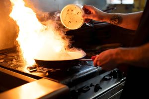 Chef flambéing food in a pan on a gas stove with dramatic flames and smoke in a professional kitchen.